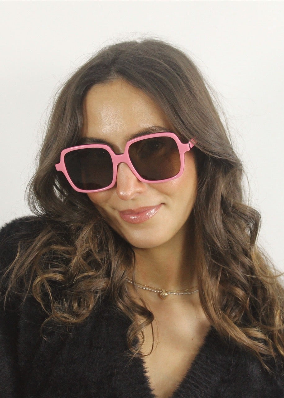 Woman wearing pink sunglasses and a black coat against a white background