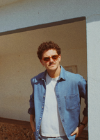 Man wearing sunglasses and a denim jacket standing in front of a building with 'AREIA' written on it.